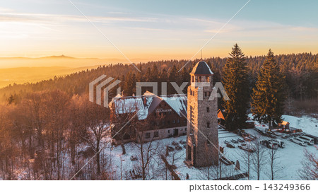 Bramberk lookout tower stands in winter with snow covering the ground. The tower is surrounded by trees and a building nearby. It is a quiet time in the late afternoon. 143249366
