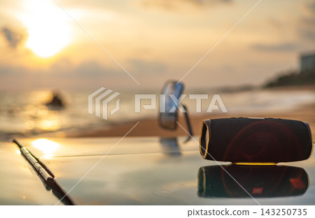 Portable Bluetooth speaker resting on the hood of a vehicle at a beach during golden hour, warm sunset light reflecting across the surface Portable Bluetooth speaker resting on the hood of a vehicle at a beach during golden hour, warm sunset light reflecting across the surface 143250735
