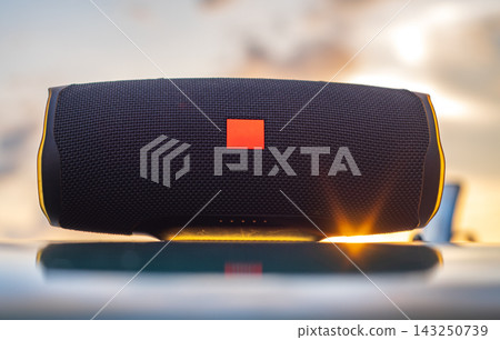 Portable Bluetooth speaker resting on the hood of a vehicle at a beach during golden hour, warm sunset light reflecting across the surface Portable Bluetooth speaker resting on the hood of a vehicle at a beach during golden hour, warm sunset light reflecting across the surface 143250739