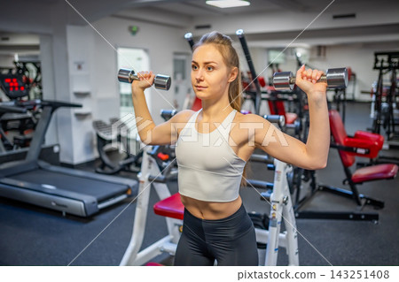 Young woman holds dumbbells in her hands and works out in the gym performing an exercise Young woman holds dumbbells in her hands and works out in the gym performing an exercise 143251408