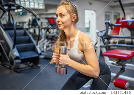 Young woman holds dumbbells in her hands and works out in the gym performing an exercise Young woman holds dumbbells in her hands and works out in the gym performing an exercise 143251426