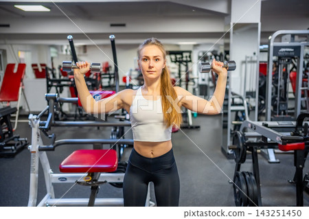 Young woman holds dumbbells in her hands and works out in the gym performing an exercise Young woman holds dumbbells in her hands and works out in the gym performing an exercise 143251450