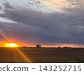 Stormy sunset illuminating a rural landscape with dramatic dark clouds, showcasing a vibrant orange sun ray breaking through the horizon over a vast open field 143252715