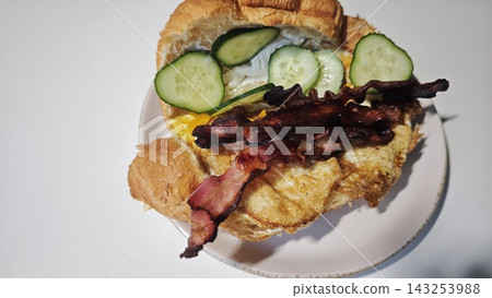 Croissant sandwich filled with fried egg, crispy bacon, sliced cucumbers, and cheddar cheese on a white plate against a plain background Croissant sandwich filled with fried egg, crispy bacon, sliced cucumbers, and cheddar cheese on a white plate against a plain background 143253988