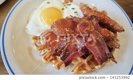 Breakfast plate featuring golden waffles topped with crispy bacon, fried egg, and sausage links, served on a white plate with blue rim Breakfast plate featuring golden waffles topped with crispy bacon, fried egg, and sausage links, served on a white plate with blue rim 143253992