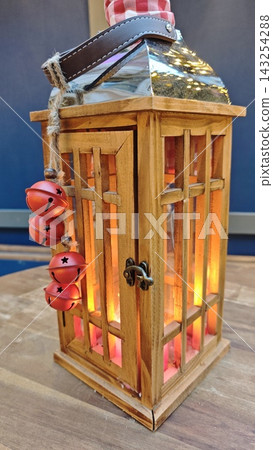 Wooden lantern with glass panels and red bells, illuminated from within, placed on a wooden surface against a blue background in a cozy indoor setting 143254288