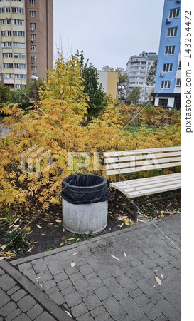 Urban park scene featuring a wooden bench beside a concrete trash can surrounded by yellow foliage and residential buildings in a city environment Urban park scene featuring a wooden bench beside a concrete trash can surrounded by yellow foliage and residential buildings in a city environment 143254472
