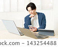 A man in his 20s working on multiple tasks, surrounded by a laptop and a tablet. 143254488