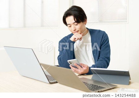 一位二十多歲的男子正在處理多項任務,周圍擺放著筆記型電腦和平板電腦。 一位二十多歲的男子正在處理多項任務,周圍擺放著筆記型電腦和平板電腦。 143254488
