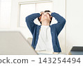 A young man sits in front of his computer, holding his head in his hands and looking up at the sky. 143254494