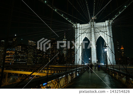 People walking along the Brooklyn Bridge pedestrian walkway with illuminated Manhattan skyline at night. Urban architecture scene with suspension cables and glowing city skyscrapers. People walking along the Brooklyn Bridge pedestrian walkway with illuminated Manhattan skyline at night. Urban architecture scene with suspension cables and glowing city skyscrapers. 143254826