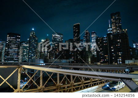 Illuminated skyscrapers of Lower Manhattan rise behind the steel roadway of the Brooklyn Bridge at night. Bright city lights and modern architecture create a dramatic New York City skyline. Illuminated skyscrapers of Lower Manhattan rise behind the steel roadway of the Brooklyn Bridge at night. Bright city lights and modern architecture create a dramatic New York City skyline. 143254856