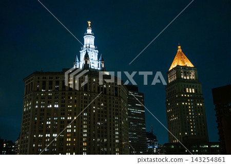 Historic towers and skyscrapers glow with warm lights against a deep blue night sky in Lower Manhattan. The illuminated skyline highlights the classic architecture of New York City. Historic towers and skyscrapers glow with warm lights against a deep blue night sky in Lower Manhattan. The illuminated skyline highlights the classic architecture of New York City. 143254861