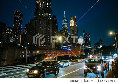 Heavy traffic moves through a Manhattan roadway under glowing streetlights and tall illuminated skyscrapers. The bright city skyline captures the vibrant nightlife of New York City. Heavy traffic moves through a Manhattan roadway under glowing streetlights and tall illuminated skyscrapers. The bright city skyline captures the vibrant nightlife of New York City. 143254864