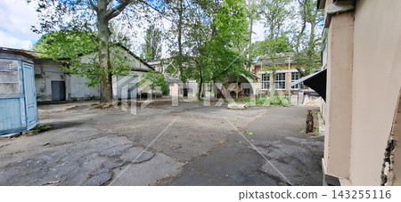 Odessa Film Studio. Old abandoned industrial courtyard with weathered buildings cracked asphalt surface and green trees on background. Odessa Film Studio. Old abandoned industrial courtyard with weathered buildings cracked asphalt surface and green trees on background. 143255116