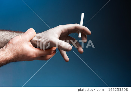 Pale crooked male hand, close-up, holding a cigarette. Healthy hand trying to quit smoking. Dark blue background. The concept of World No Tobacco Day Pale crooked male hand, close-up, holding a cigarette. Healthy hand trying to quit smoking. Dark blue background. The concept of World No Tobacco Day 143255208