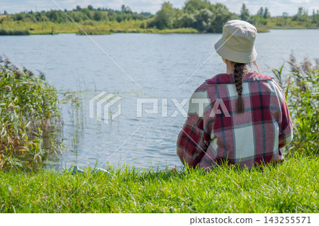 Woman in a light sun hat and checked shirt sitting on a grassy hill facing a wide lake on a clear day. Lakeside leisure and peaceful connection with nature 143255571