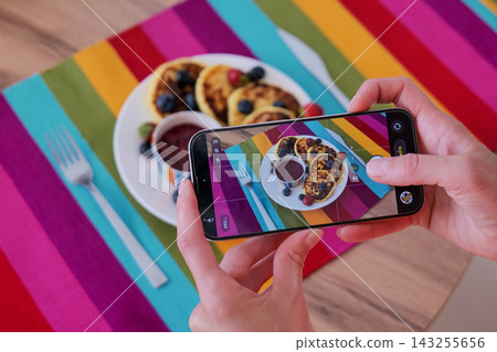 Hands hold a smartphone horizontally while photographing pancakes with sauce and cutlery. Mobile food photography supports digital content creation Hands hold a smartphone horizontally while photographing pancakes with sauce and cutlery. Mobile food photography supports digital content creation 143255656