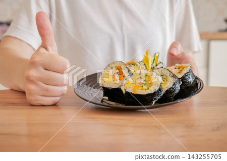 Plate of sliced Korean kimbap with hand giving thumbs up in background. Korean food approval and successful homemade meal concept Plate of sliced Korean kimbap with hand giving thumbs up in background. Korean food approval and successful homemade meal concept 143255705