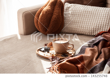 Cozy warm hot chocolate with marshmallows and chocolate squares on a tray next to a blanket and pillows indoors 143255748