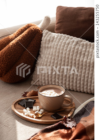 Cozy hot chocolate with marshmallows and chocolate squares on a wooden tray near plush pillows and a checkered blanket 143255750