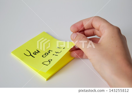 A hand lifts the corner of a yellow sticky note with a handwritten You can do it message from a stack on a white surface. Motivational reminder concept 143255812