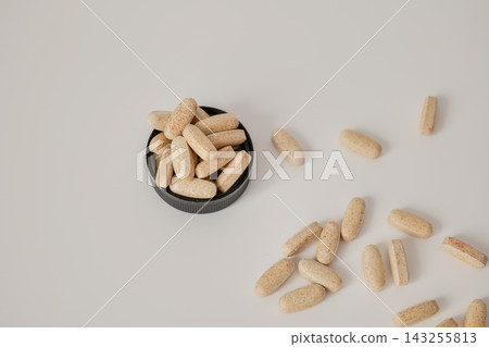 Beige capsules filling a black round lid with loose capsules scattered on a white surface. Capsule quantity and supplement organization concept Beige capsules filling a black round lid with loose capsules scattered on a white surface. Capsule quantity and supplement organization concept 143255813