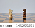Light and dark chess knights face each other on a glossy wooden board. The minimal duel represents competition, confrontation, and strategic opposition 143255851