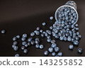 An overturned plastic cup in the upper right spills blueberries into a narrowing trail across a dark surface. Diagonal composition creates copy space for harvest, food styling, and abundance concepts. 143255852