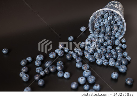 An overturned plastic cup in the upper right spills blueberries into a narrowing trail across a dark surface. Diagonal composition creates copy space for harvest, food styling, and abundance concepts. An overturned plastic cup in the upper right spills blueberries into a narrowing trail across a dark surface. Diagonal composition creates copy space for harvest, food styling, and abundance concepts. 143255852