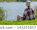 Woman in a light sun hat and checked shirt sitting beside a lake with her legs near the water and looking toward the opposite shore. Lakeside relaxation and nature escape 143255857