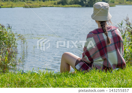 Woman in a light sun hat and checked shirt sitting beside a lake with her legs near the water and looking toward the opposite shore. Lakeside relaxation and nature escape Woman in a light sun hat and checked shirt sitting beside a lake with her legs near the water and looking toward the opposite shore. Lakeside relaxation and nature escape 143255857
