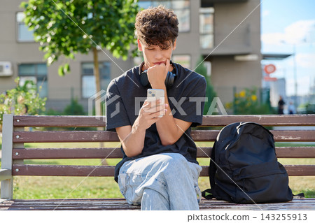 Thoughtful young man using smartphone sitting park bench outdoors Thoughtful young man using smartphone sitting park bench outdoors 143255913