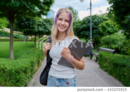 Smiling teenage girl holding laptop and backpack standing in park portrait Smiling teenage girl holding laptop and backpack standing in park portrait 143255915