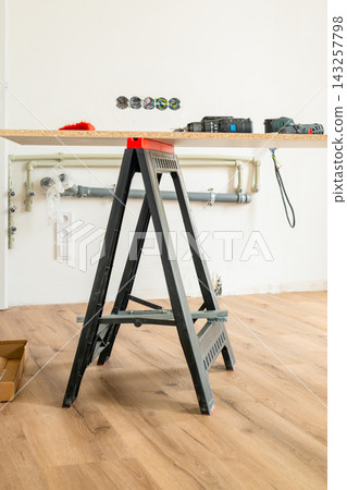 Constructing a temporary work surface using a black folding ladder and a wooden board in a home setting. Visible tools include drills and electrical components arranged on the countertop. Constructing a temporary work surface using a black folding ladder and a wooden board in a home setting. Visible tools include drills and electrical components arranged on the countertop. 143257798