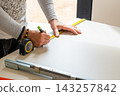 Measuring and marking a wood panel using a tape measure on a clean workbench. The workspace features tools such as a pencil and metal ruler. 143257842
