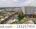 Elevated panoramic view of a dense urban neighborhood with apartment towers, low-rise buildings, busy streets, and Vietnamese flags beneath a cloudy sky. 143257955