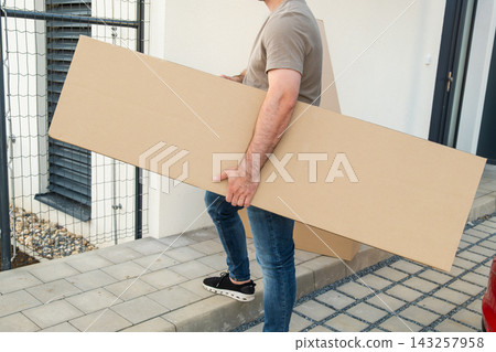 Man transporting a large cardboard panel while moving along a sidewalk outside a residential building. Man transporting a large cardboard panel while moving along a sidewalk outside a residential building. 143257958