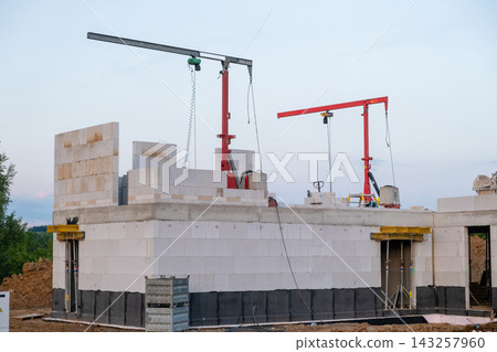 Construction cranes positioned above a residential building site, lifting heavy block materials. The structure features concrete walls and an organized workspace with construction materials visible. Construction cranes positioned above a residential building site, lifting heavy block materials. The structure features concrete walls and an organized workspace with construction materials visible. 143257960