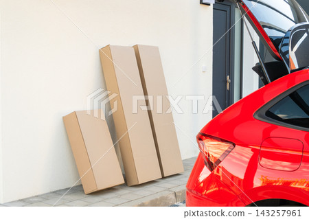 Large cardboard delivery boxes positioned against a wall next to a parked red vehicle. Clear sky and modern residential setting visible in the background. Large cardboard delivery boxes positioned against a wall next to a parked red vehicle. Clear sky and modern residential setting visible in the background. 143257961