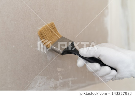 A hand wearing a white glove applies finishing paint to a smooth drywall surface using a high quality brush. 143257983