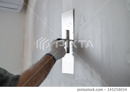 Finishing contractor using stainless steel trowel to apply a smooth layer of plaster on interior wall. Clean workspace with bright lighting and minimal distractions visible in background. Finishing contractor using stainless steel trowel to apply a smooth layer of plaster on interior wall. Clean workspace with bright lighting and minimal distractions visible in background. 143258034