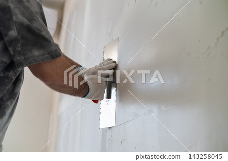 Skilled technician smoothing finishing compound with a stainless steel trowel on drywall during home renovation. Clean workspace environment with partially finished walls visible in the background. Skilled technician smoothing finishing compound with a stainless steel trowel on drywall during home renovation. Clean workspace environment with partially finished walls visible in the background. 143258045