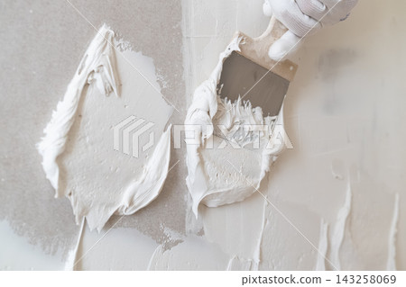 Applying white plaster using a stainless steel spatula on a smooth wall surface. The workspace features a clean and organized environment with subtle gray tones. Applying white plaster using a stainless steel spatula on a smooth wall surface. The workspace features a clean and organized environment with subtle gray tones. 143258069