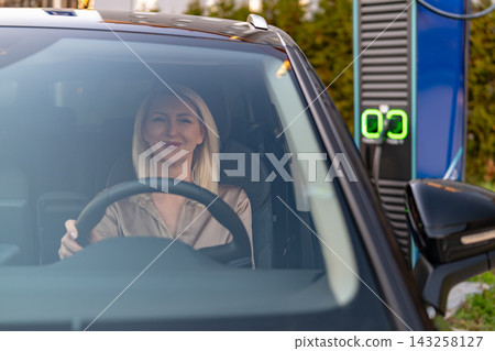 A woman is seated inside an electric vehicle, preparing to charge at a contemporary EV charging station. A woman is seated inside an electric vehicle, preparing to charge at a contemporary EV charging station. 143258127