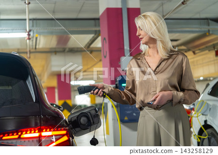 A woman connects an electric vehicle to a charging station in a contemporary parking garage. The environment features brightly lit spaces and multiple charging points for various vehicles. A woman connects an electric vehicle to a charging station in a contemporary parking garage. The environment features brightly lit spaces and multiple charging points for various vehicles. 143258129