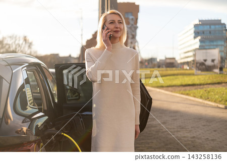 Business professional communicating via mobile phone next to electric vehicle in modern urban environment. Clean, contemporary architecture and greenery visible in the background. Business professional communicating via mobile phone next to electric vehicle in modern urban environment. Clean, contemporary architecture and greenery visible in the background. 143258136