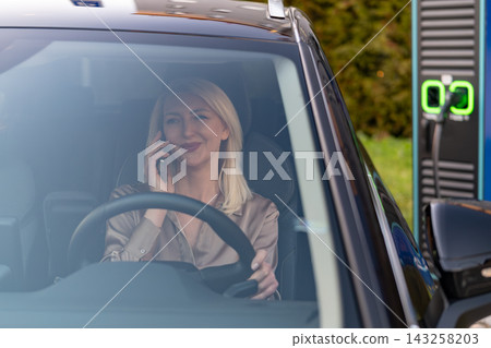 Blonde female driver using mobile phone while parked at electric vehicle charging station. Cleanly designed charging unit and green indicator light visible in the background, showcasing an eco 143258203