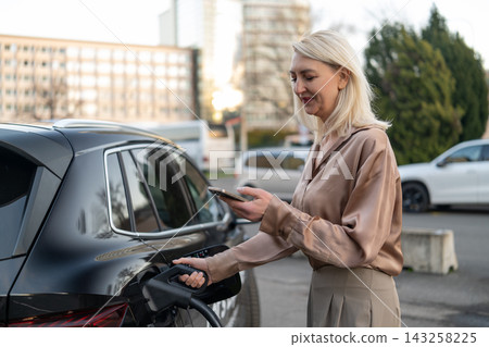 Driver engages in electric vehicle charging while interacting with mobile application at urban charging station. The setting features modern buildings and greenery visible in the background. Driver engages in electric vehicle charging while interacting with mobile application at urban charging station. The setting features modern buildings and greenery visible in the background. 143258225