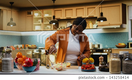 Chef vlogger using lemon juice and olive oil for seasoning the salad during filming, easy homemade recipe under natural light. Black woman showcasing meal prep and culinary development. Chef vlogger using lemon juice and olive oil for seasoning the salad during filming, easy homemade recipe under natural light. Black woman showcasing meal prep and culinary development. 143258414